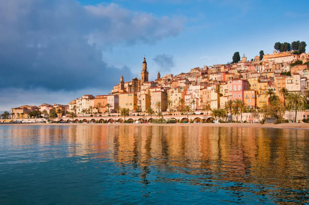 Colorful hillside village of Menton in Provence reflected in calm water, with pastel buildings and palm-lined waterfront.