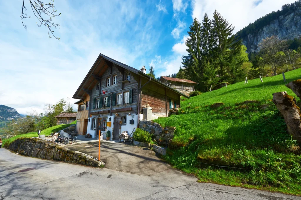 Traditional Swiss alpine chalet set on a green hillside with forested mountains and clear blue sky in the background.