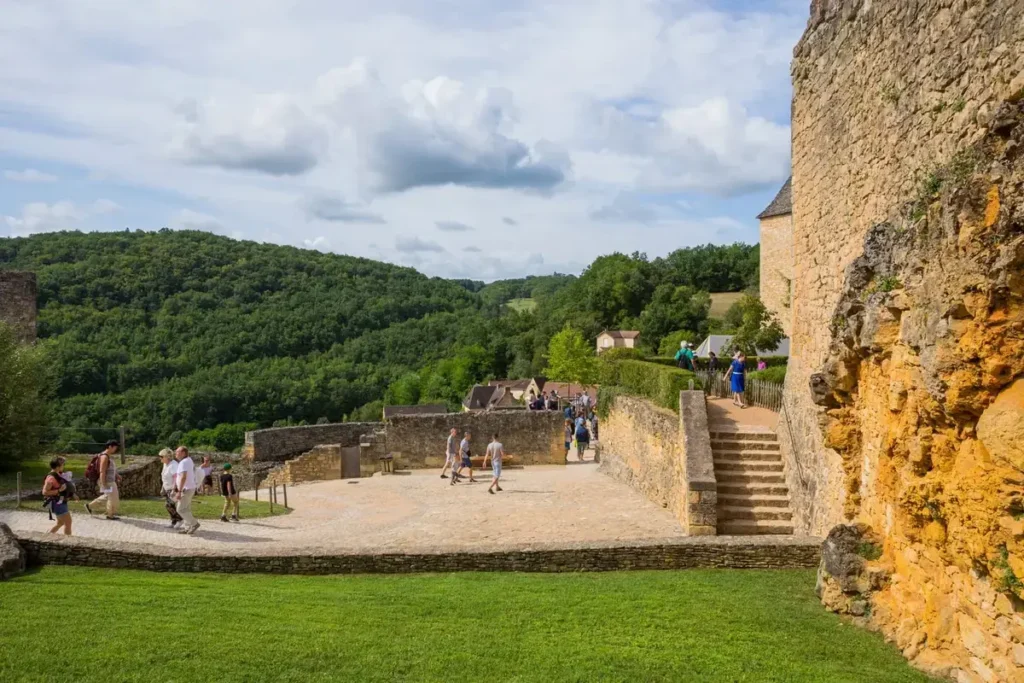 Visitors walking through a stone castle terrace with steps, walls, and forested Dordogne hills in the background