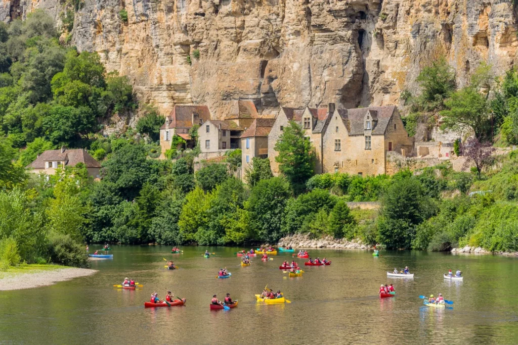 Colorful canoes on Dordogne River passing cliffside village and limestone cliffs during scenic canoe trip