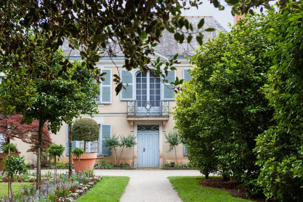 French mansion with pale blue shutters, flowering garden beds, trimmed greenery, and a gravel path leading to the entrance.