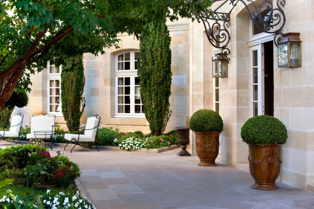 Elegant French mansion terrace with stone walls, tall windows, potted topiary, garden seating, and manicured planting.