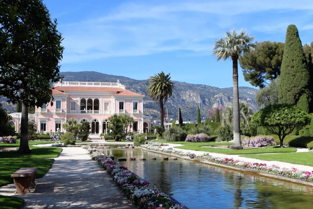 Pink French Riviera mansion with formal gardens, reflecting pool, palm trees, flowers, and mountain views.