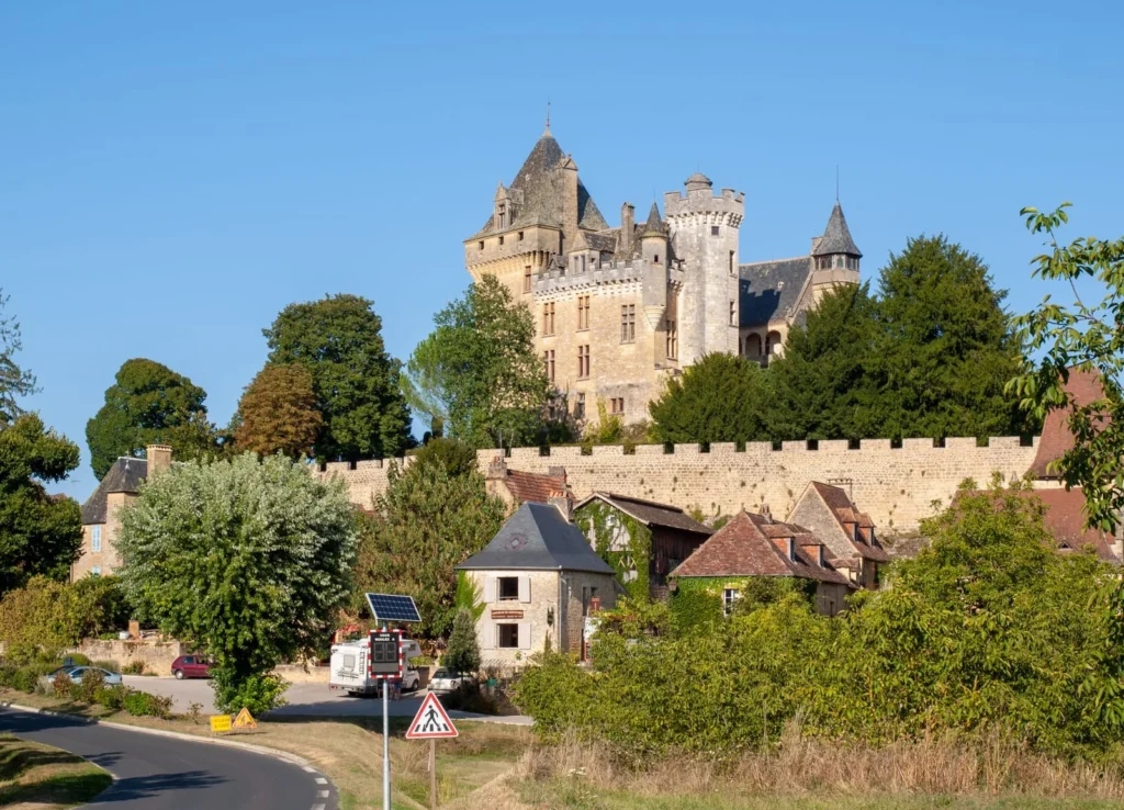 Stone château rising above village rooftops and a winding road in the Dordogne countryside.