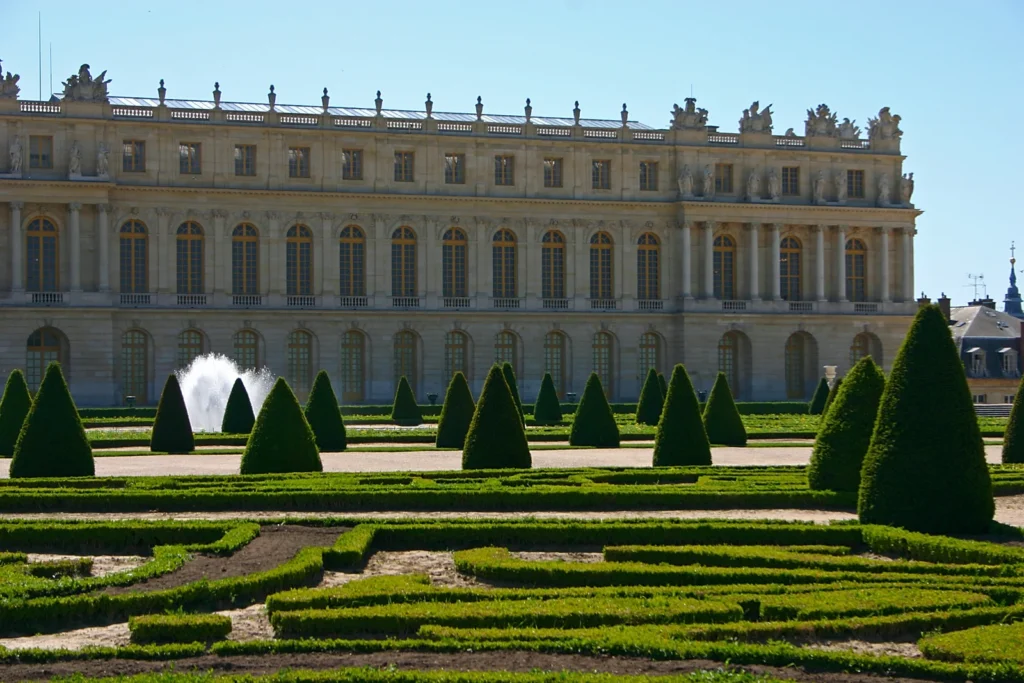 Palace of Versailles facade with formal gardens, clipped hedges, and a fountain in the foreground.