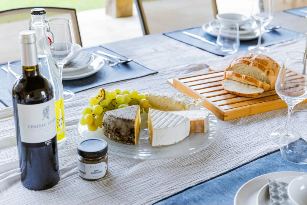 Gourmet dining table with wine, cheese platter, grapes, bread, and flowers in a rustic stone interior.