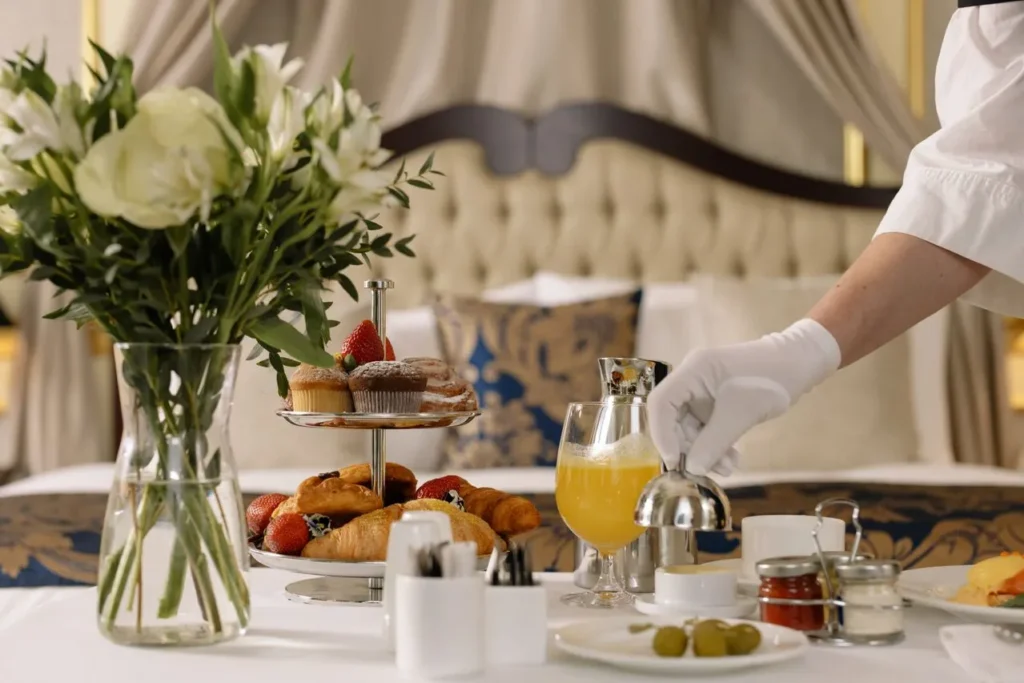 A waiter serving fresh orange juice and pastries on a refined breakfast table with flowers, croissants, and fruits in a luxury room setting