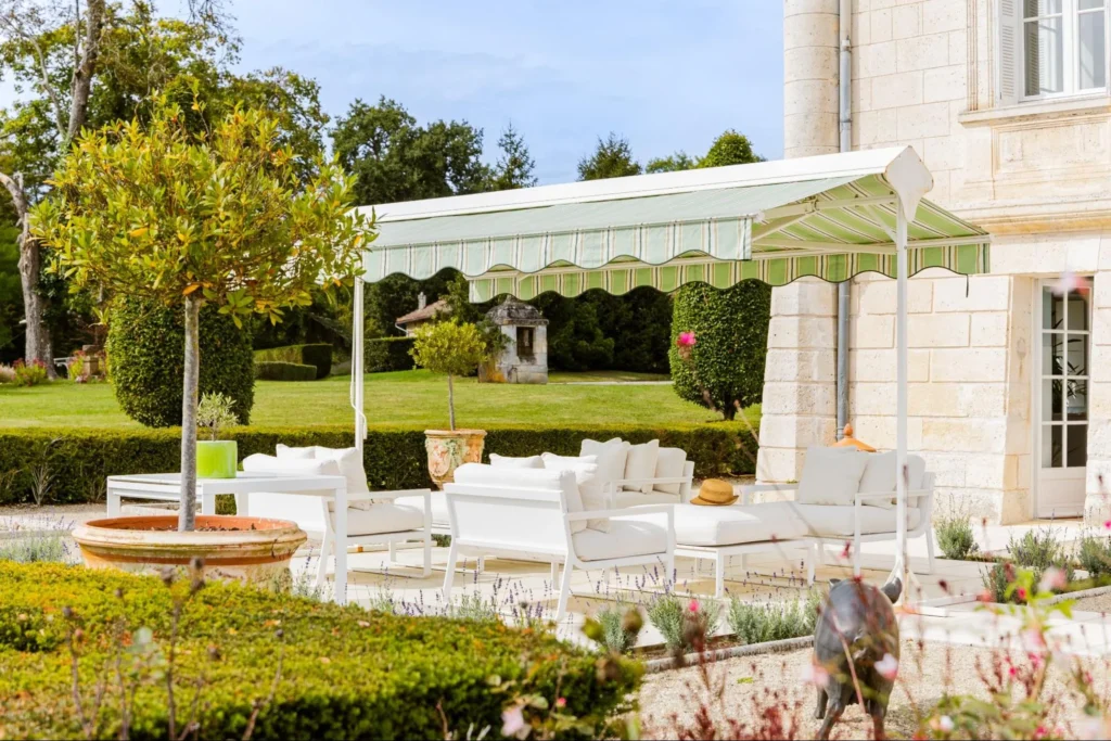 Elegant outdoor lounge with white seating under a canopy in a landscaped garden beside a stone building.