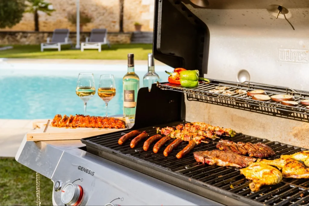 Outdoor barbecue grill with meats, vegetables, and wine glasses beside a swimming pool in a luxury villa setting.