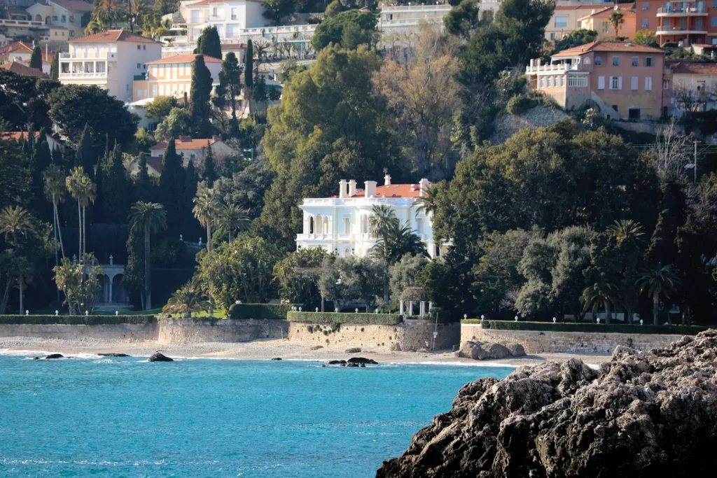 Seaside villa overlooking turquoise water, rocky coast, palm trees, and hillside homes.