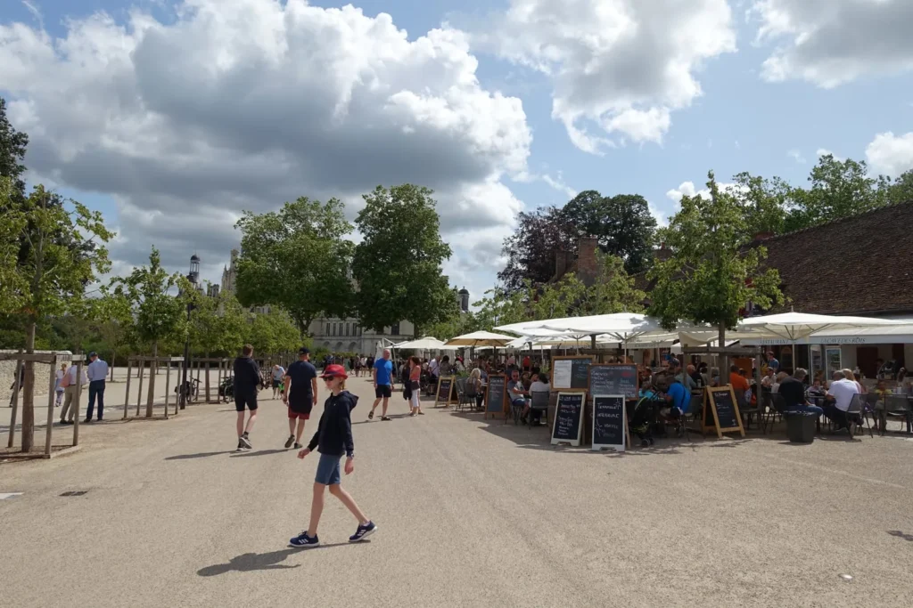 Outdoor cafe square with visitors, restaurant terraces, trees, and a chateau in the distance