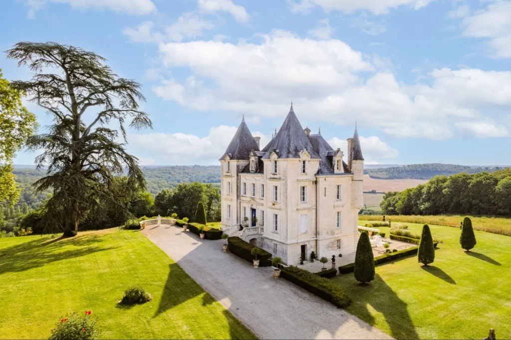 Grand French chateau surrounded by lawns, trees, and countryside views under a blue sky